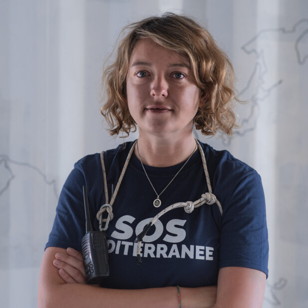 A white woman with wavy blonde hair wearing a navy blue t-shirt with a stethoscope around her neck