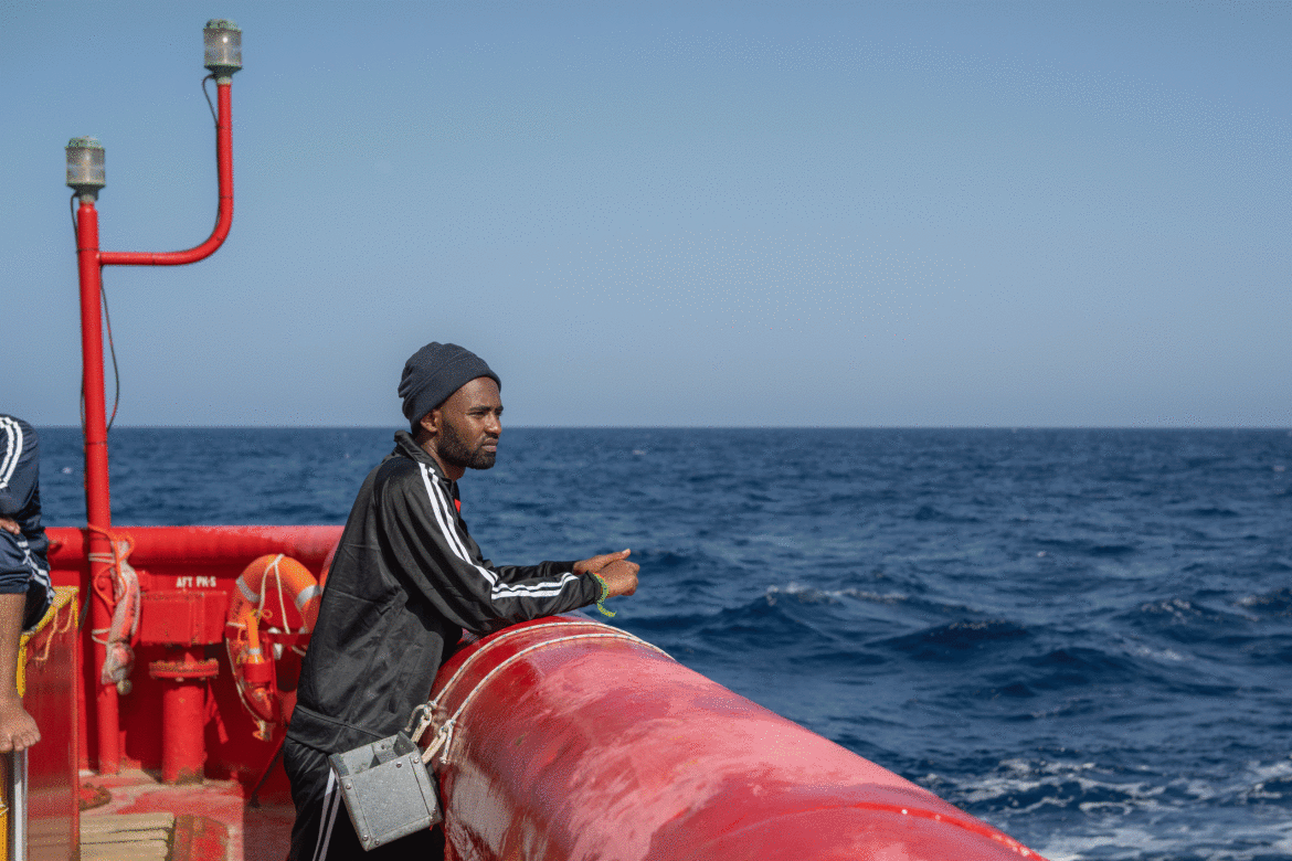 A man stands at the edge of a ship, looking out over the open sea on a clear day.