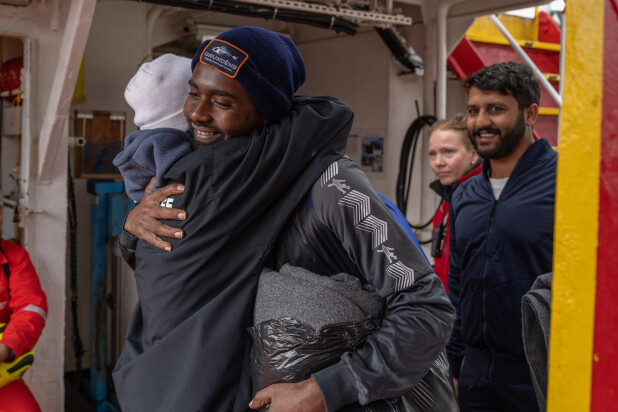Two people embrace warmly on a ship while others nearby look on and smile.