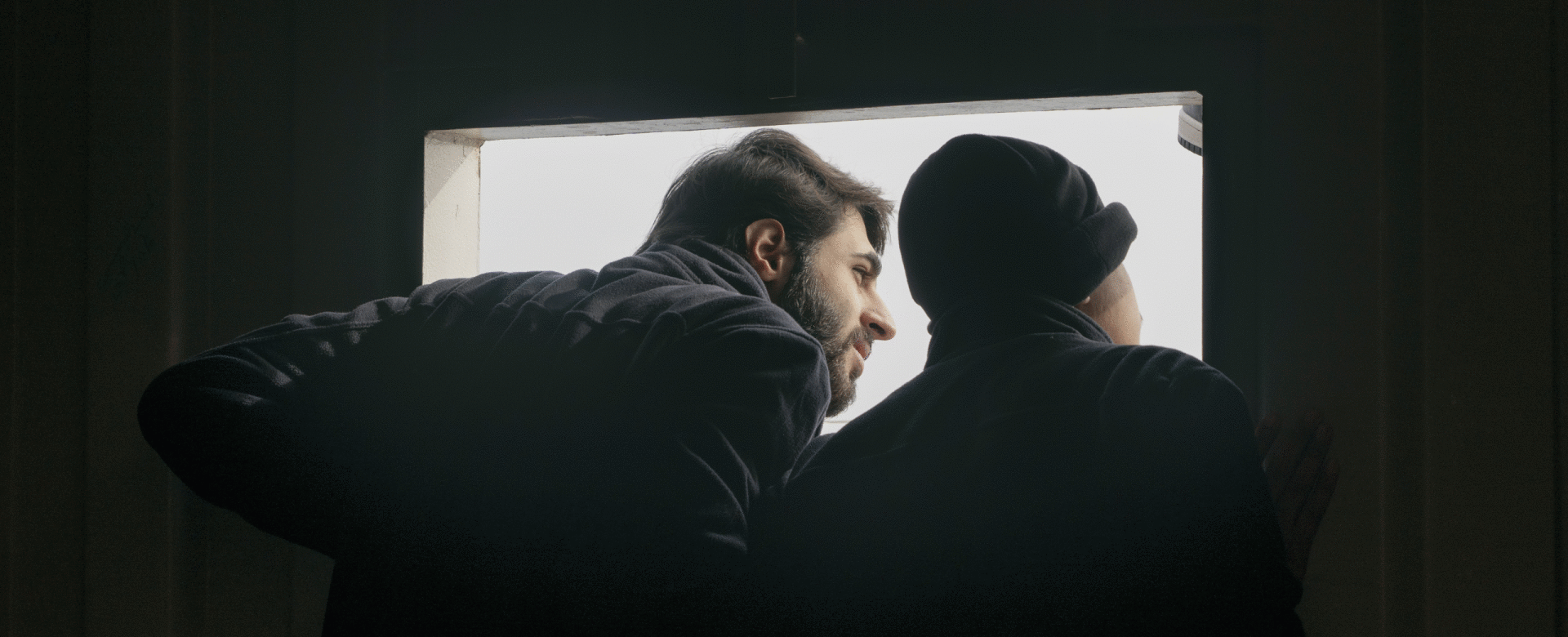 Two people lean through a window-like opening on a ship, looking outside toward the light.