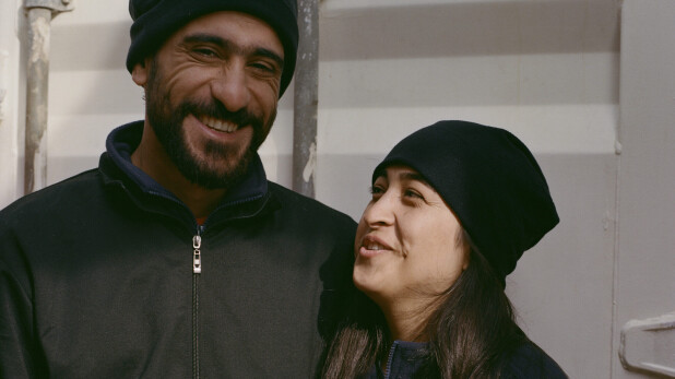 A smiling couple in dark jackets and hats stands close together on a ship’s deck.