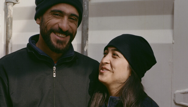 A smiling couple in dark jackets and hats stands close together on a ship’s deck.