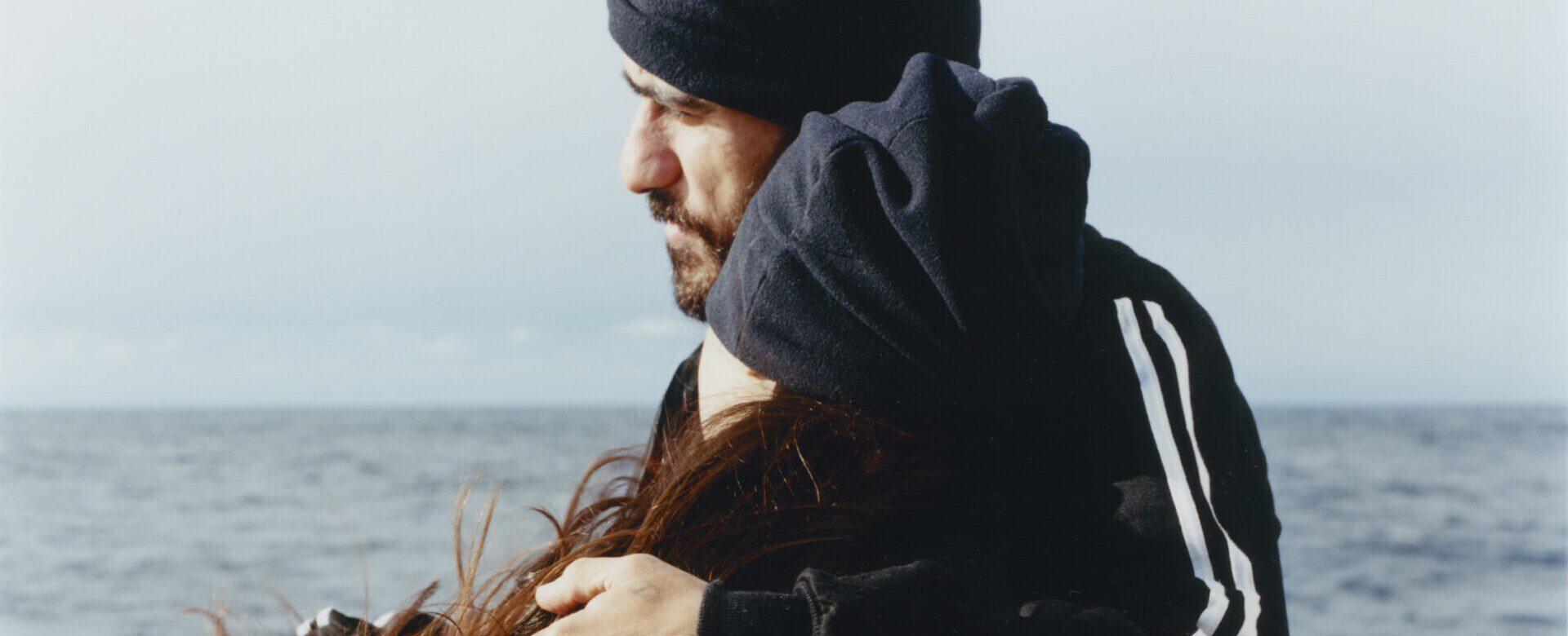 A man embraces a woman on a ship’s deck, both wearing dark hats and facing the open sea.