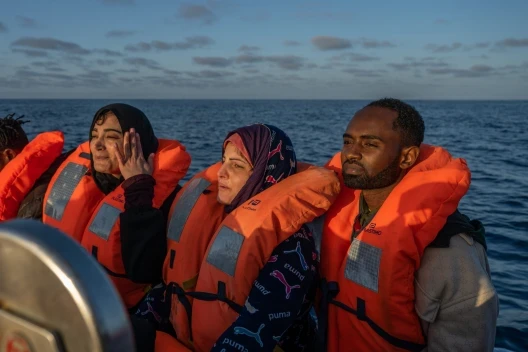 Three people in bright orange life jackets sit closely together on a rescue boat at sea.