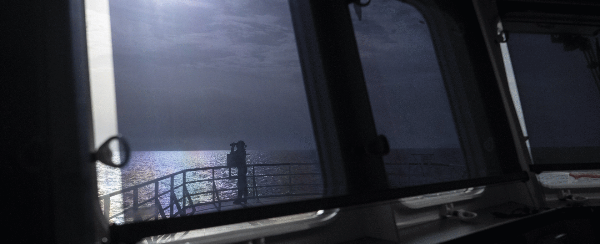 A dark ship interior looking out through large windows at the sea under moonlight. The ocean and sh