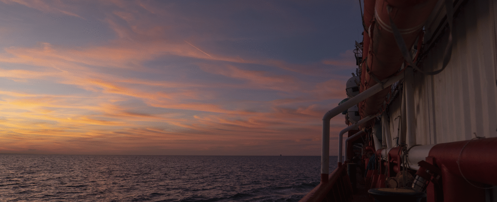 Sunset over the sea viewed from a ship’s deck, with soft clouds and orange light in the sky.