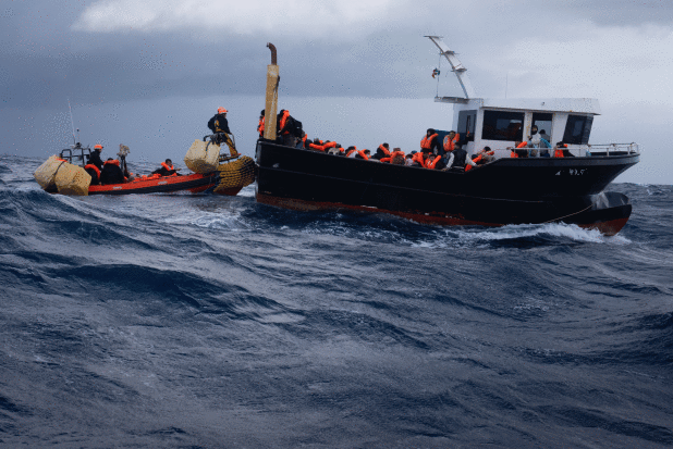 Two overcrowded boats move through rough sea waters under a cloudy sky. Dozens of people wearing or
