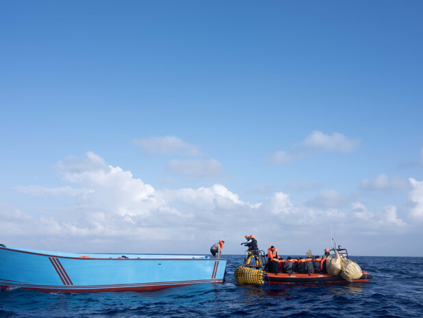 Rescue crew in a small inflatable boat approaches a large blue wooden boat in open sea.