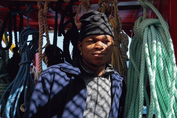 A man stands among hanging ropes on a ship, with shadows cast across his face and jacket.