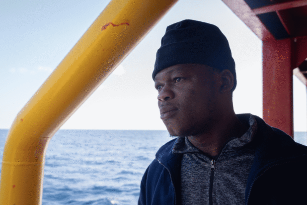 A man in a beanie looks out at the sea from a ship’s deck beside a yellow support beam.