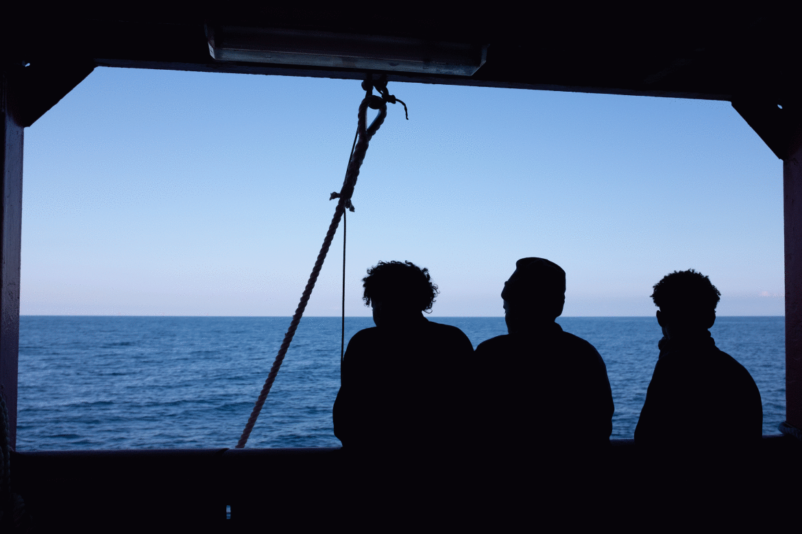 Three people in silhouette stand at the railing of a ship, looking out over a calm ocean under a cl