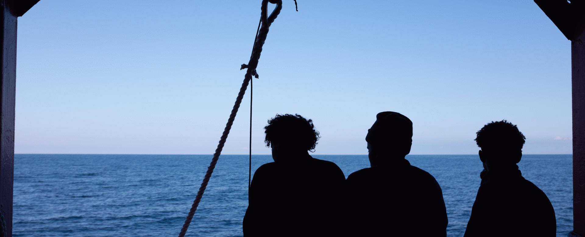 Three people in silhouette stand at the railing of a ship, looking out over a calm ocean under a cl