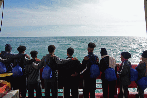 A group of young people stand arm-in-arm on a ship’s deck, facing the sea with blue bags on their b