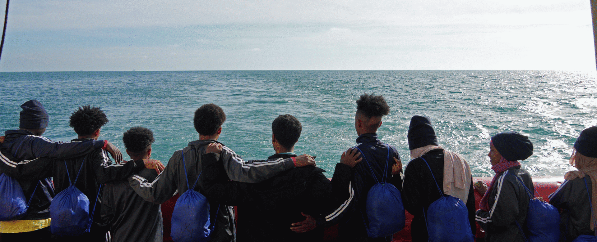A group of young people stand arm-in-arm on a ship’s deck, facing the sea with blue bags on their b